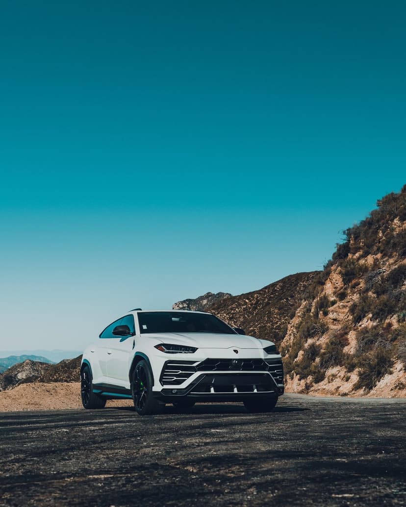 White Lamborghini Urus SUV parked on a gravel mountain road under a bright blue sky with rugged peaks behind it.