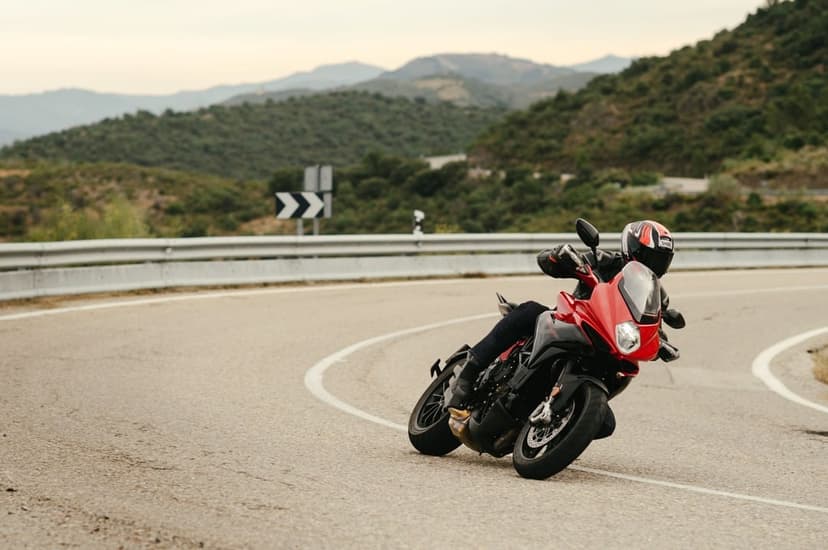 Red motorcycle leaning into a sharp turn on a winding mountain road with green hills in the background.