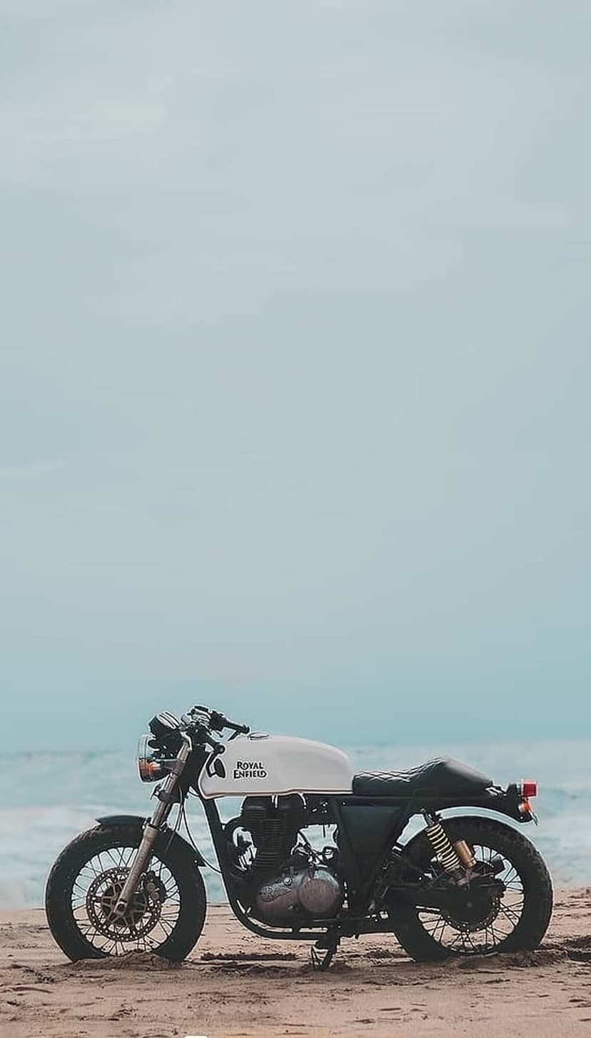White Royal Enfield motorcycle parked on a sandy beach with soft ocean waves under a clear blue sky.