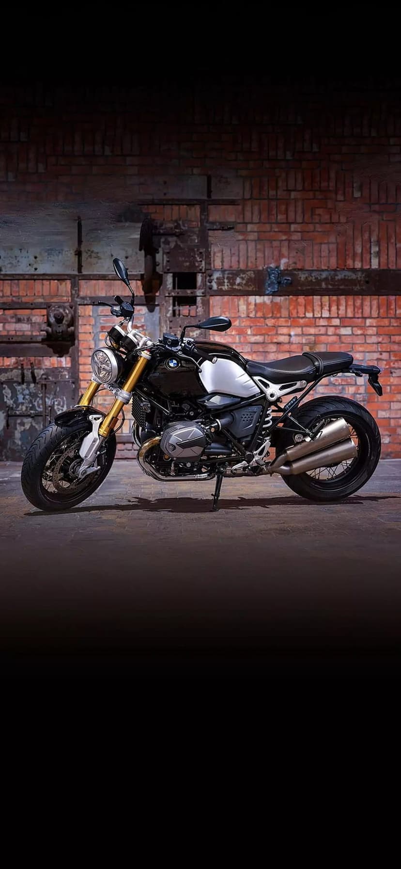 BMW R nineT motorcycle featuring gold forks and a silver tank parked against a weathered industrial brick wall.