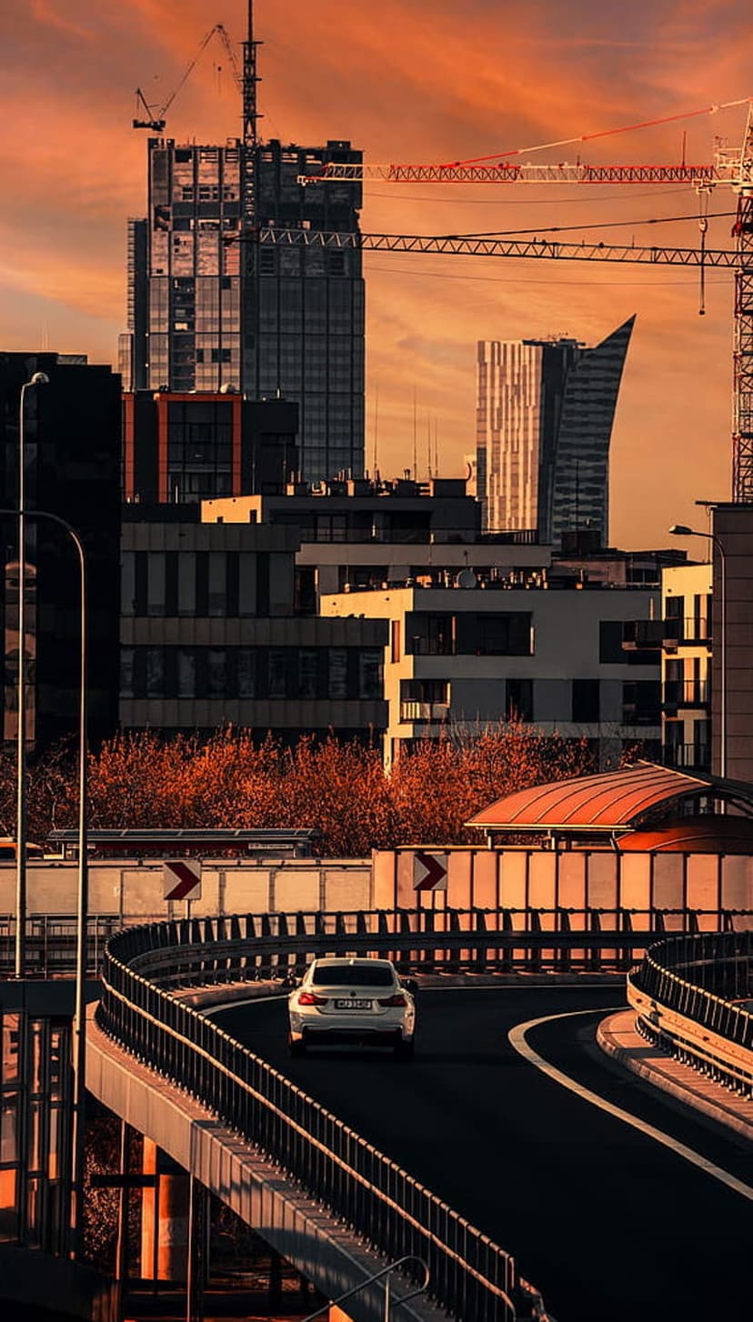 White car driving on a curving urban highway toward tall skyscrapers and construction cranes under a sunset sky.
