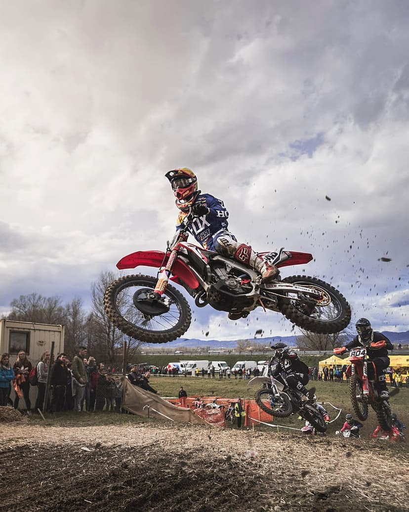 Motocross rider on a red and white bike jumps mid-air over a dirt track with spectators under a cloudy sky.