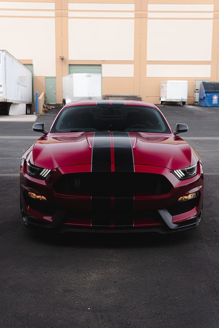 Red Ford Mustang GT with black hood stripes parked on asphalt in front of industrial buildings and dumpsters.