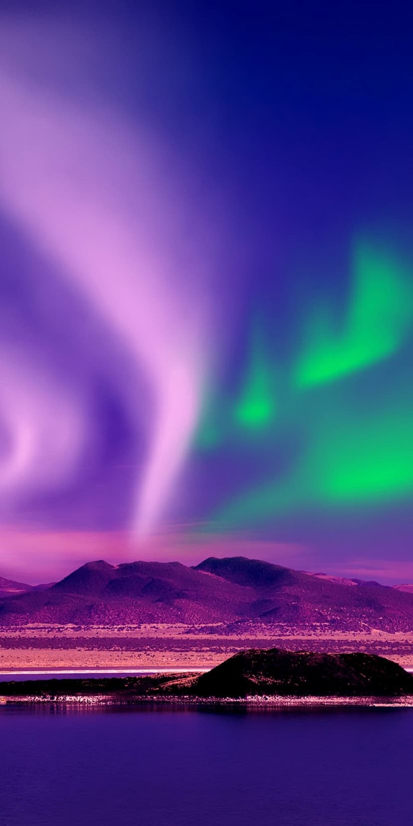Purple and green northern lights dance above a jagged mountain range and reflect in a calm alpine lake at night.