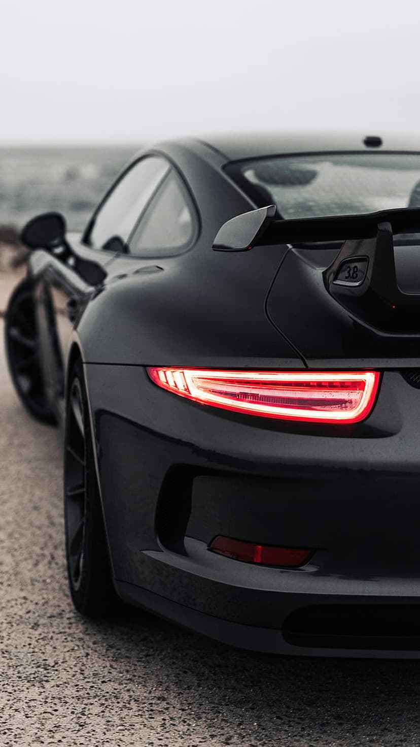 Porsche 911 GT3 rear profile with glowing red taillights and wing on a gravel coastal road at twilight.