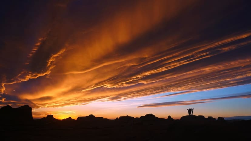 Silhouetted people stand on a rocky desert ledge under a sky filled with fiery orange and dark storm clouds.