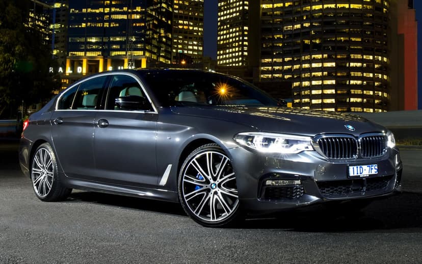Charcoal BMW 5 Series sedan with glowing headlights parked in front of illuminated city skyscrapers at night.
