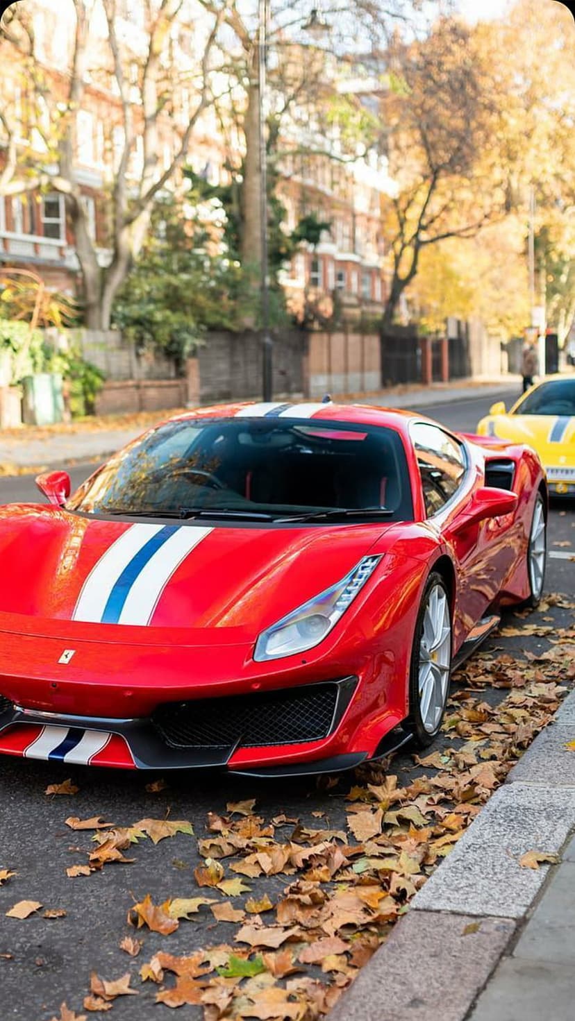 Red Ferrari with blue racing stripes parked on a street covered in orange autumn leaves with a yellow car behind.