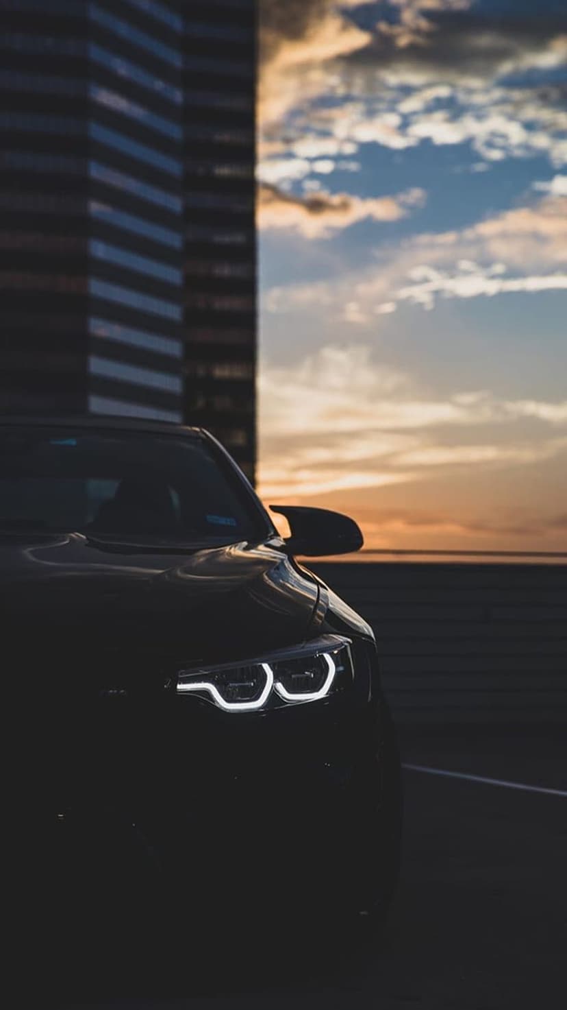 BMW LED headlights glowing in front of a tall city skyscraper under a fiery orange and purple sunset sky.