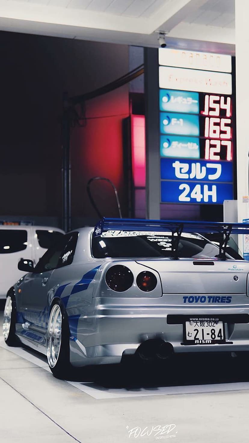 Nissan GT-R R34 with blue racing stripes parked at a glowing Japanese gas station at night under neon signs.