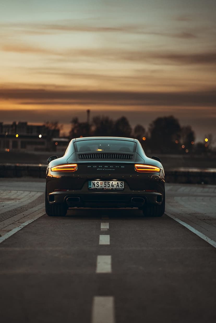 Porsche 911 Carrera rear view on an asphalt road at sunset with glowing red taillights and a cloudy orange sky.