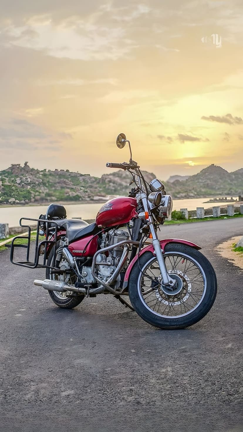 Red Royal Enfield motorcycle on a road by a lake during a golden sunset with distant hills and orange clouds.