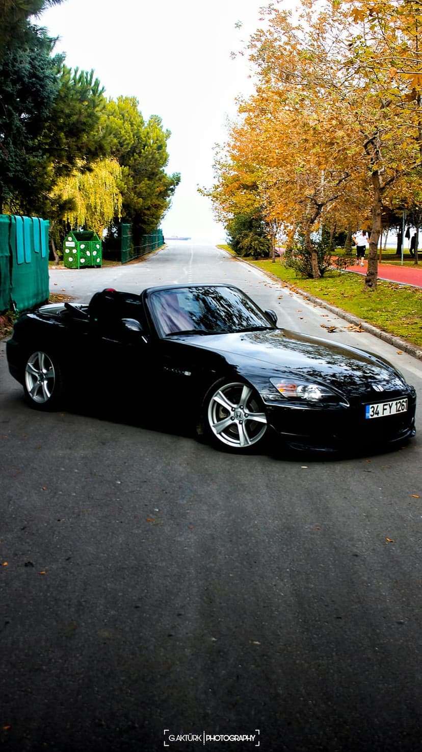 Black convertible sports car parked on a gray asphalt road surrounded by vibrant orange and red autumn forest trees.