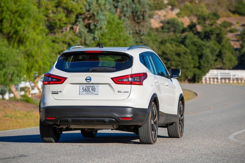 Rear view of a white Nissan Rogue Sport SL AWD driving on a winding asphalt road through green hills.