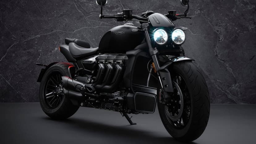 Matte black Triumph Rocket 3 R motorcycle with glowing dual headlights against a dark textured stone wall.