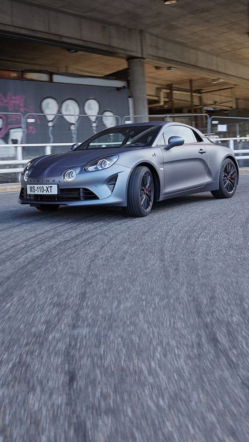 Alpine A110 sports car with red calipers drives under a concrete bridge featuring colorful graffiti art.