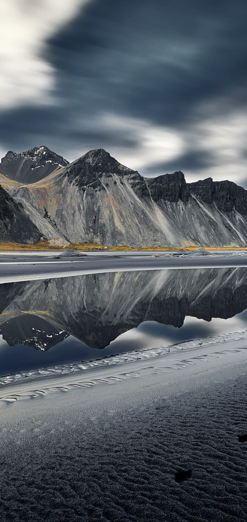 Rugged mountain peaks with snow patches reflect in a calm coastal lagoon under a streaked, dynamic sky.