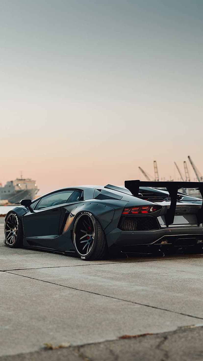 Lamborghini Aventador with a widebody kit and rear wing parked at an industrial port during a golden sunset.