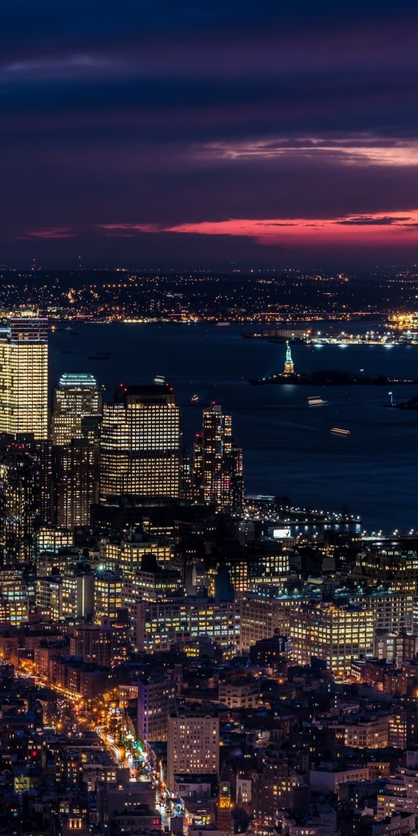 New York City skyline at night with the Statue of Liberty and glowing harbor reflections under a purple sky.