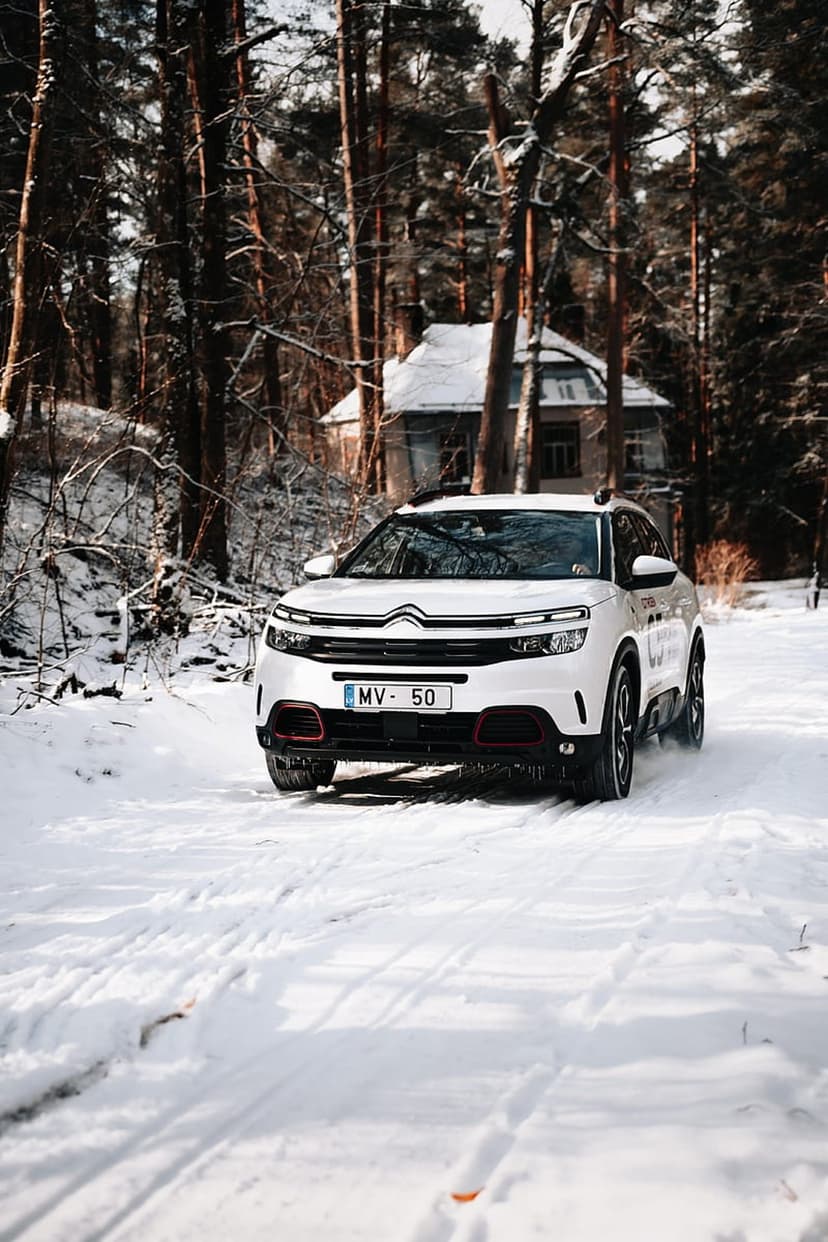 White SUV with glowing headlights drives through deep snow past a weathered house in a dense winter forest.