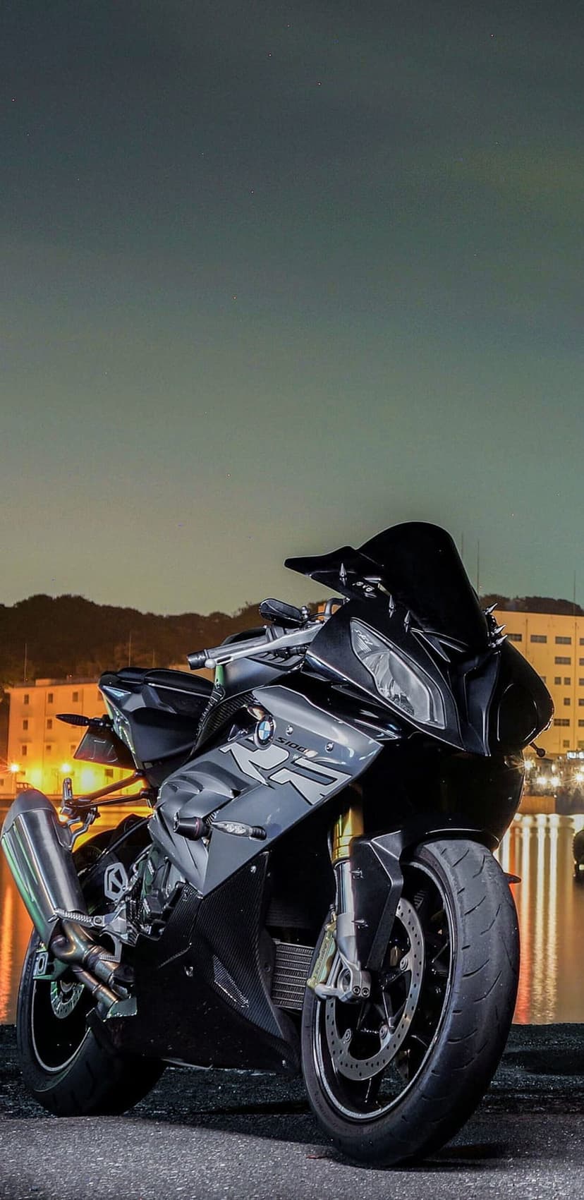 Black BMW S1000RR sportbike stands beside dark water reflecting distant city lights and hills at night.