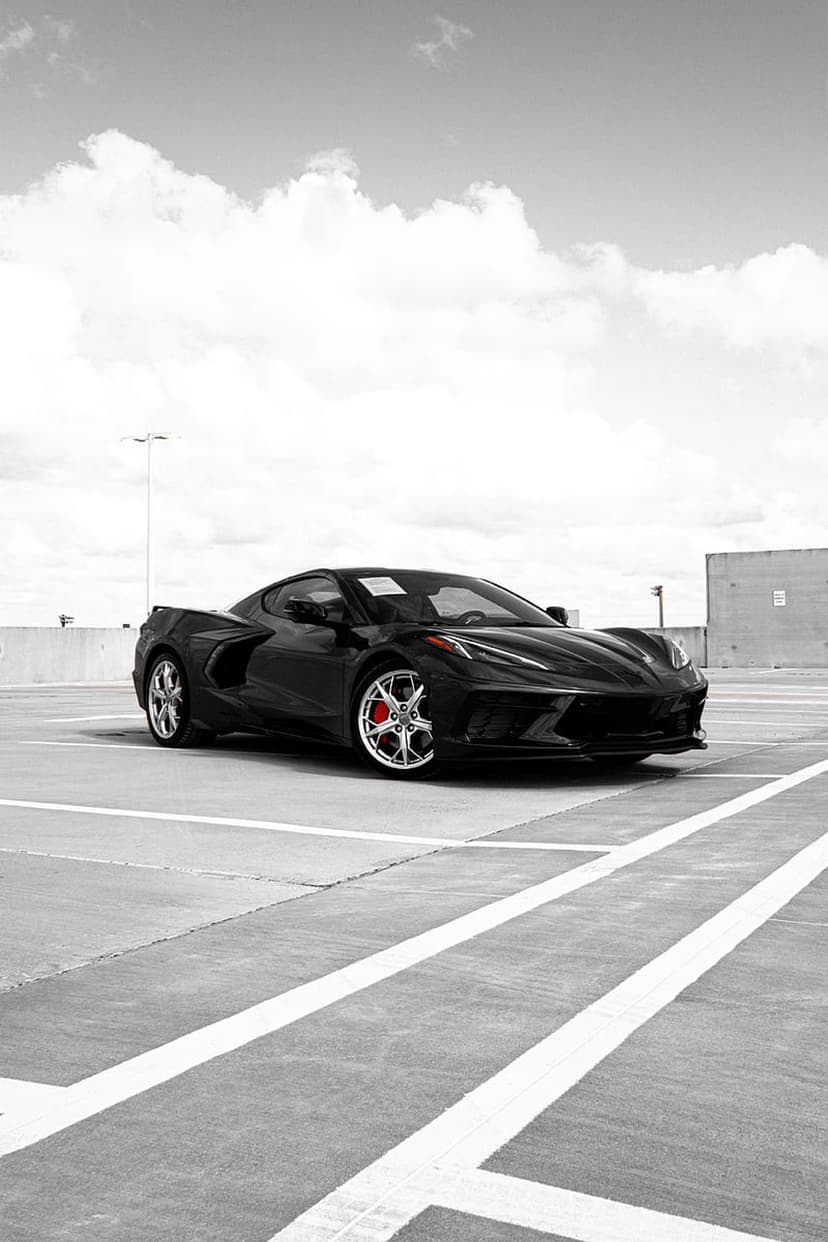 Black Chevrolet Corvette C8 parked on concrete under a bright sky with white clouds in a vertical composition.