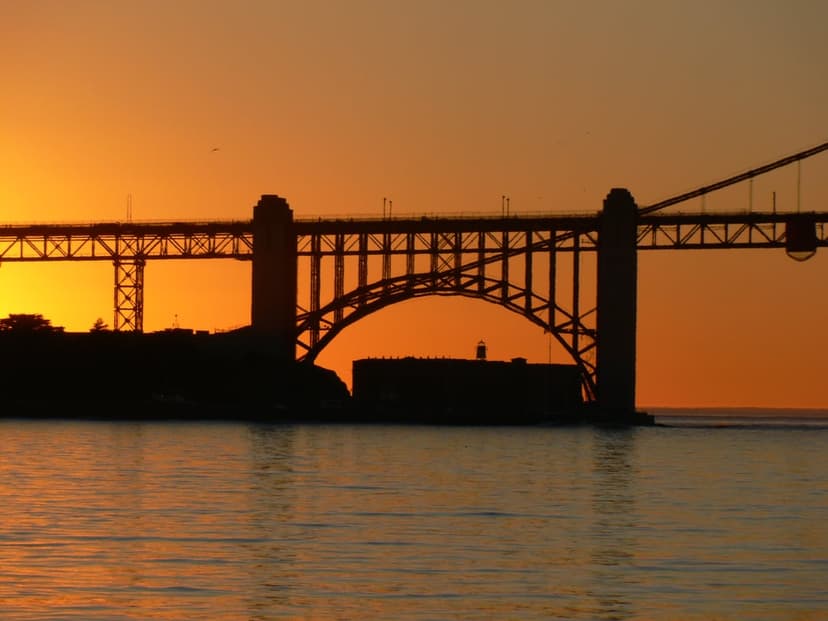 Golden Gate Bridge silhouette stands against a vibrant orange sunset with calm water reflections in the bay.