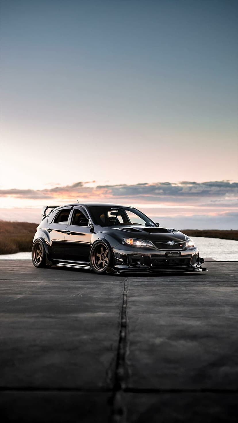 Black Subaru WRX STI hatchback with bronze wheels and a widebody kit parked on asphalt during a coastal sunset.
