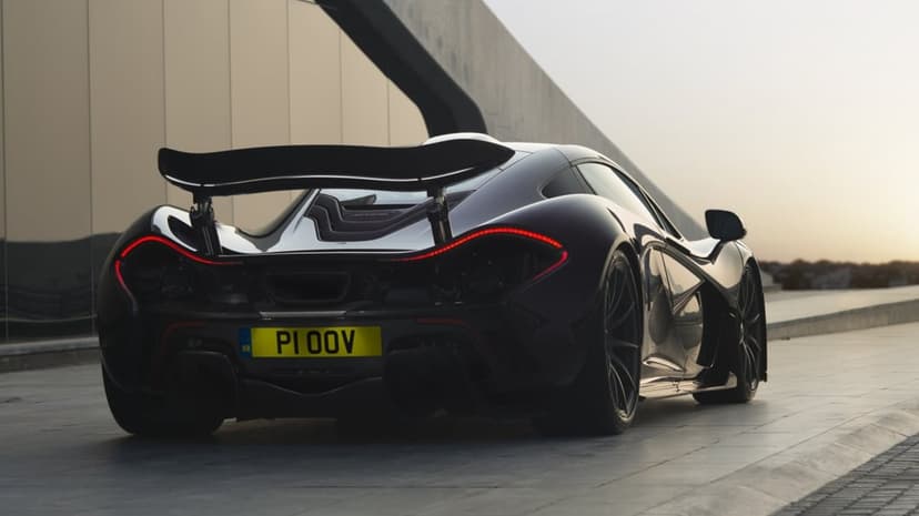 Rear view of a dark McLaren P1 hypercar with curved red LED taillights at sunset next to a modern building.