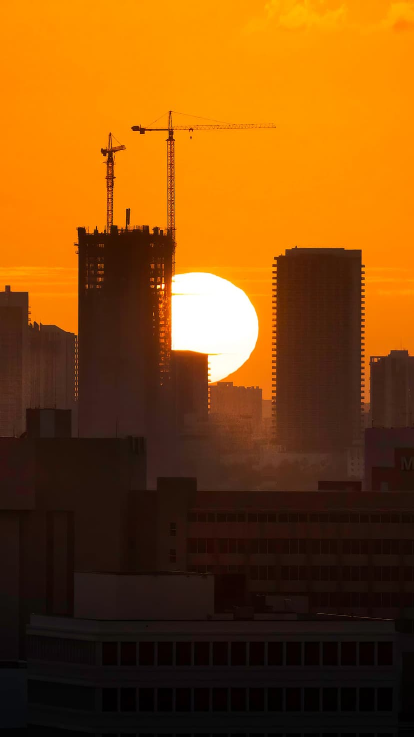 Glowing sun emerges behind a silhouetted skyscraper and construction cranes under a bright orange dawn sky.