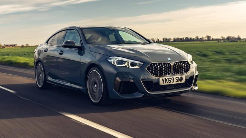 Grey BMW M235i Gran Coupe driving on a rural road with green fields under a soft afternoon sky background.