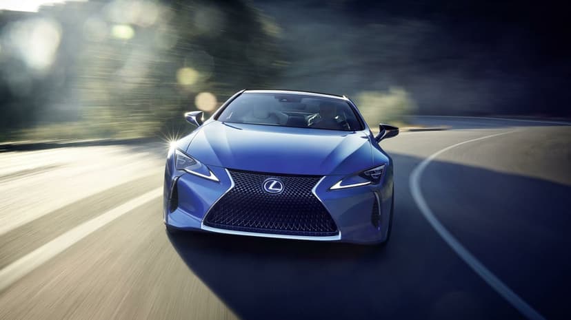 Metallic blue Lexus LC 500 coupe driving fast on a curved road with motion blur and bright sun lens flares.