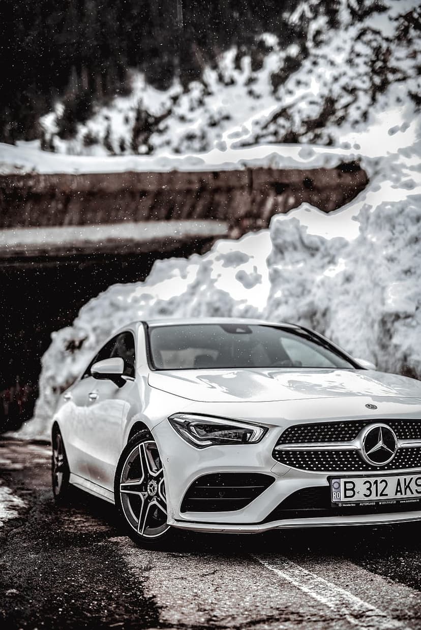 White Mercedes sedan parked on a wet mountain road between massive snowdrifts under a light snowfall.