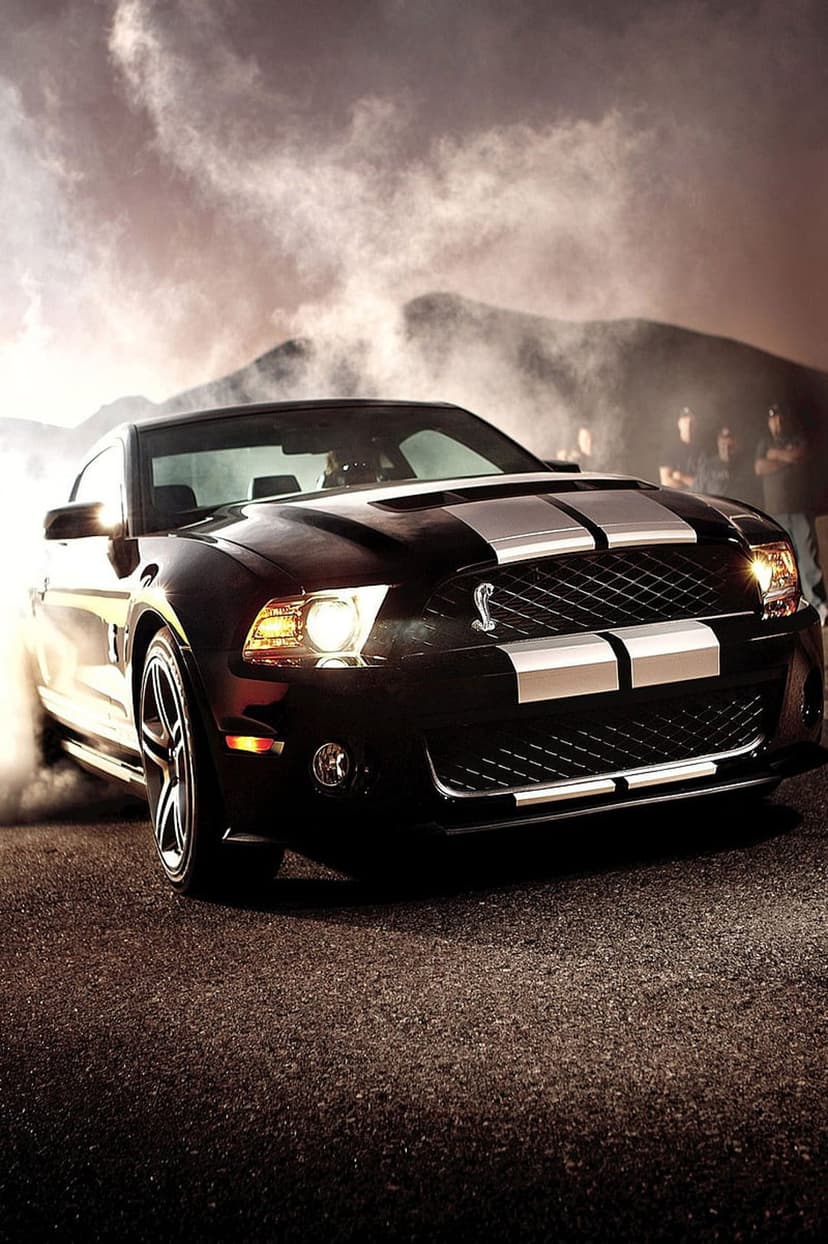 Black Ford Mustang Shelby Cobra performs a burnout with white smoke clouds against a mountain backdrop.