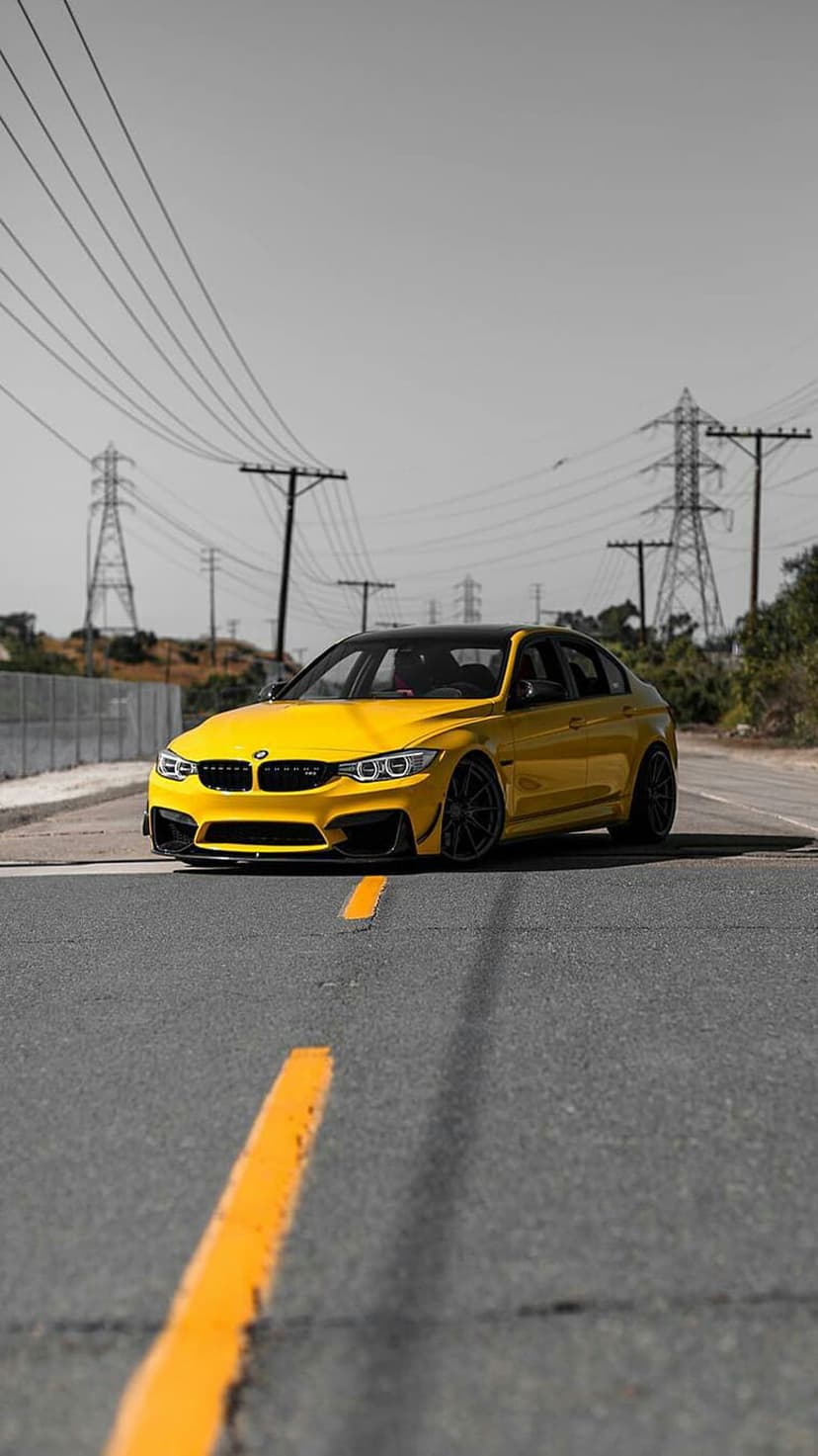 Yellow BMW M3 performance sedan with black wheels on an asphalt road beneath industrial electricity towers.