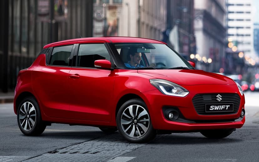 Red Suzuki Swift hatchback on a city street with a driver inside and blurred urban buildings in the background.