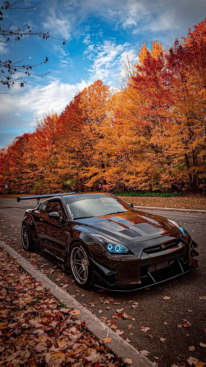Black Nissan GTR sports car with blue headlights parked on a road lined with orange and red autumn trees.