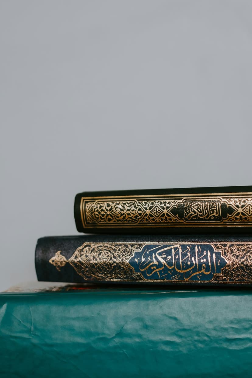 Stacked Islamic books with gold Arabic calligraphy on dark green and teal covers against a grey background.