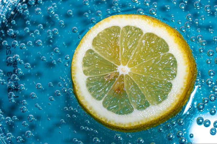 Yellow lemon slice submerged in bubbling blue liquid with small effervescent spheres clinging to the citrus pulp.