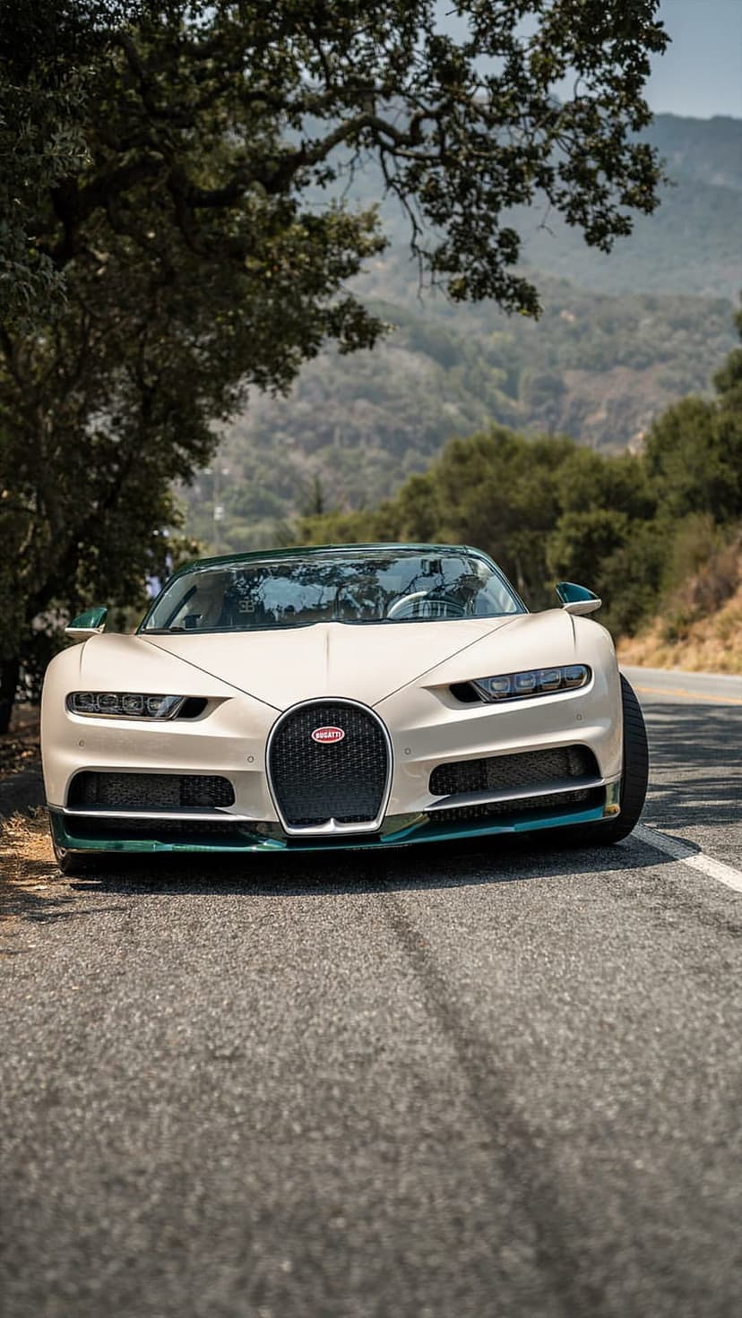 White Bugatti Chiron parked on a paved winding road surrounded by lush green trees and distant rolling hills.