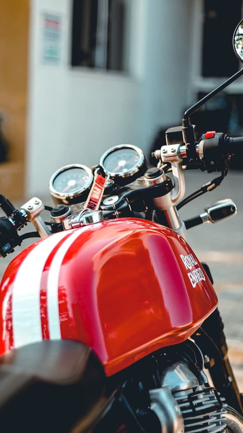 Red Royal Enfield fuel tank with white racing stripes, chrome gauges, and handlebars in a close-up motorcycle shot.