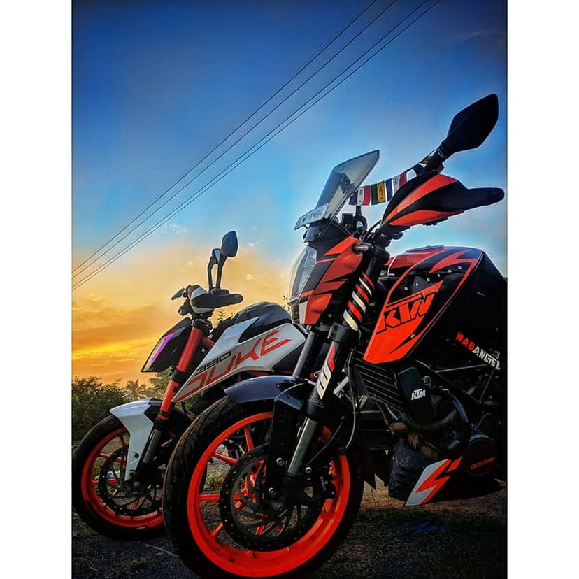 KTM Duke motorcycles stand side-by-side against a glowing orange twilight sky with overhead power cables.