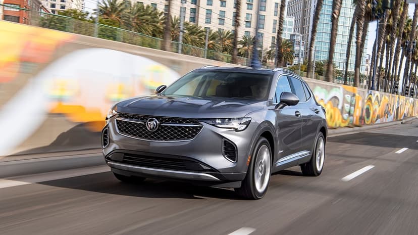 A grey Buick Envision SUV driving on a city street with motion blur, palm trees, and urban buildings.