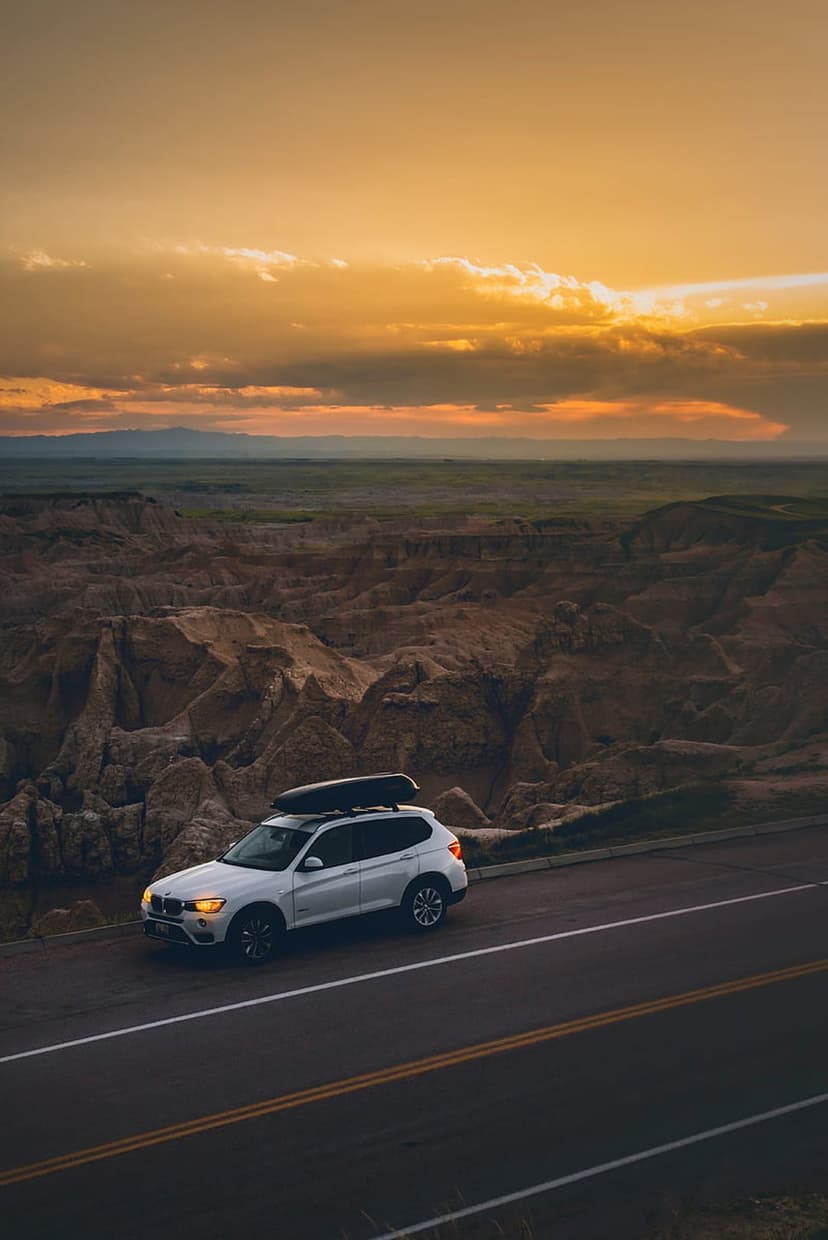 White SUV with roof rack driving on a mountain road under a vibrant orange sunset sky with golden clouds.