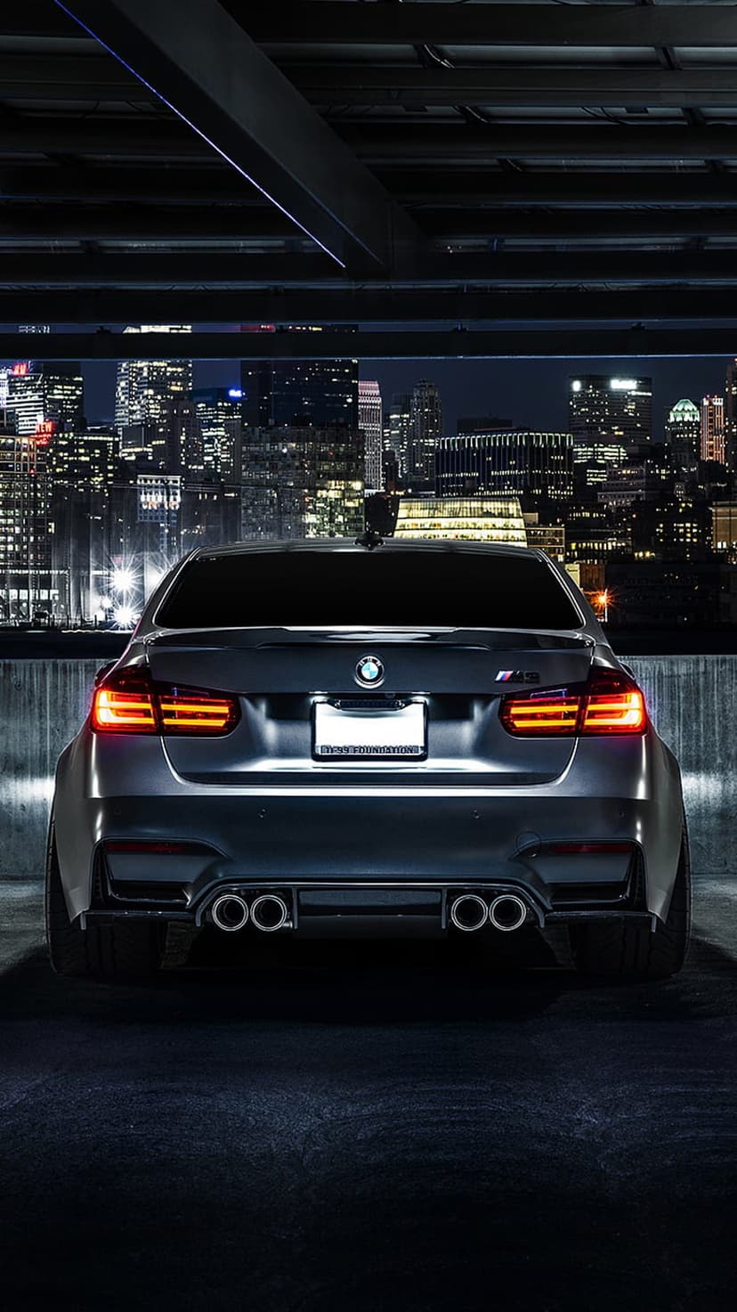 BMW M3 rear view with glowing taillights in a dark parking garage overlooking a lit metropolitan city skyline.