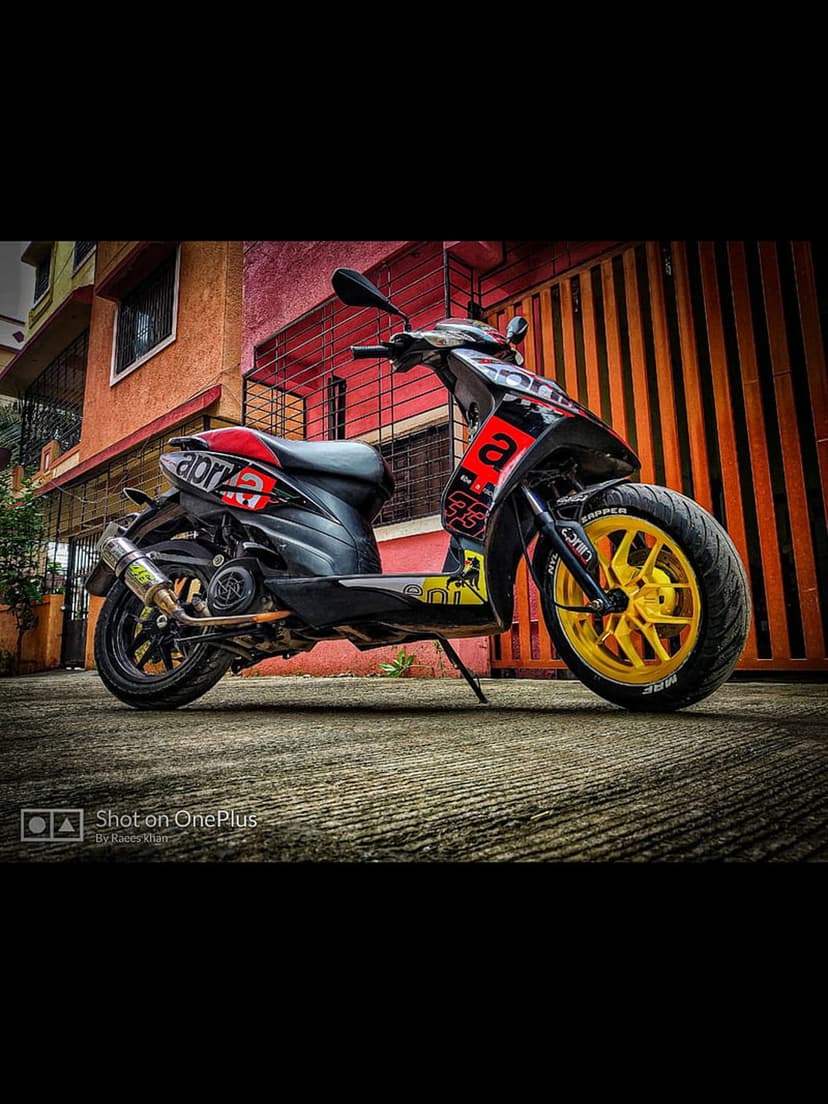 Black and red Aprilia scooter with yellow rims parked in front of an orange and blue urban building wall.