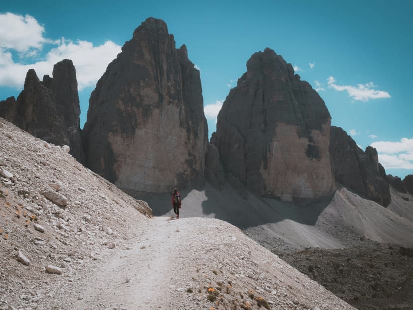 Hiker on a gravel trail walking toward the three jagged mountain peaks of Tre Cime di Lavaredo under a blue sky.