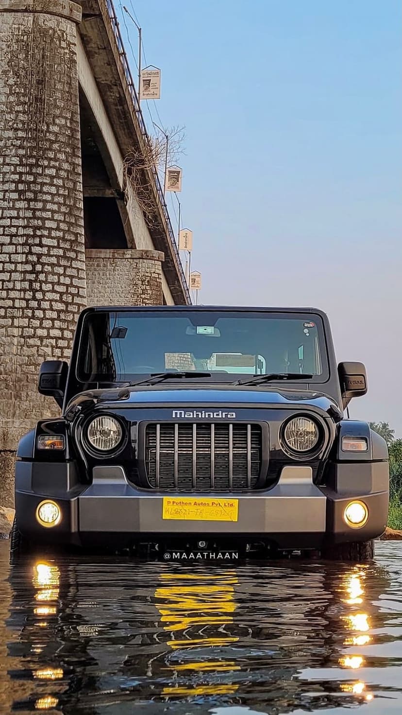 Black Mahindra Thar SUV partially submerged in a rippling river next to a weathered stone bridge under a blue sky.
