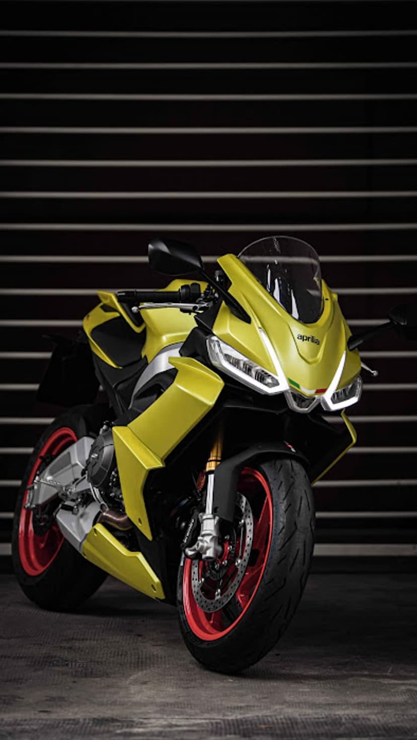 Yellow Aprilia motorcycle with red wheels and LED headlights on a grey surface against a dark striped wall.