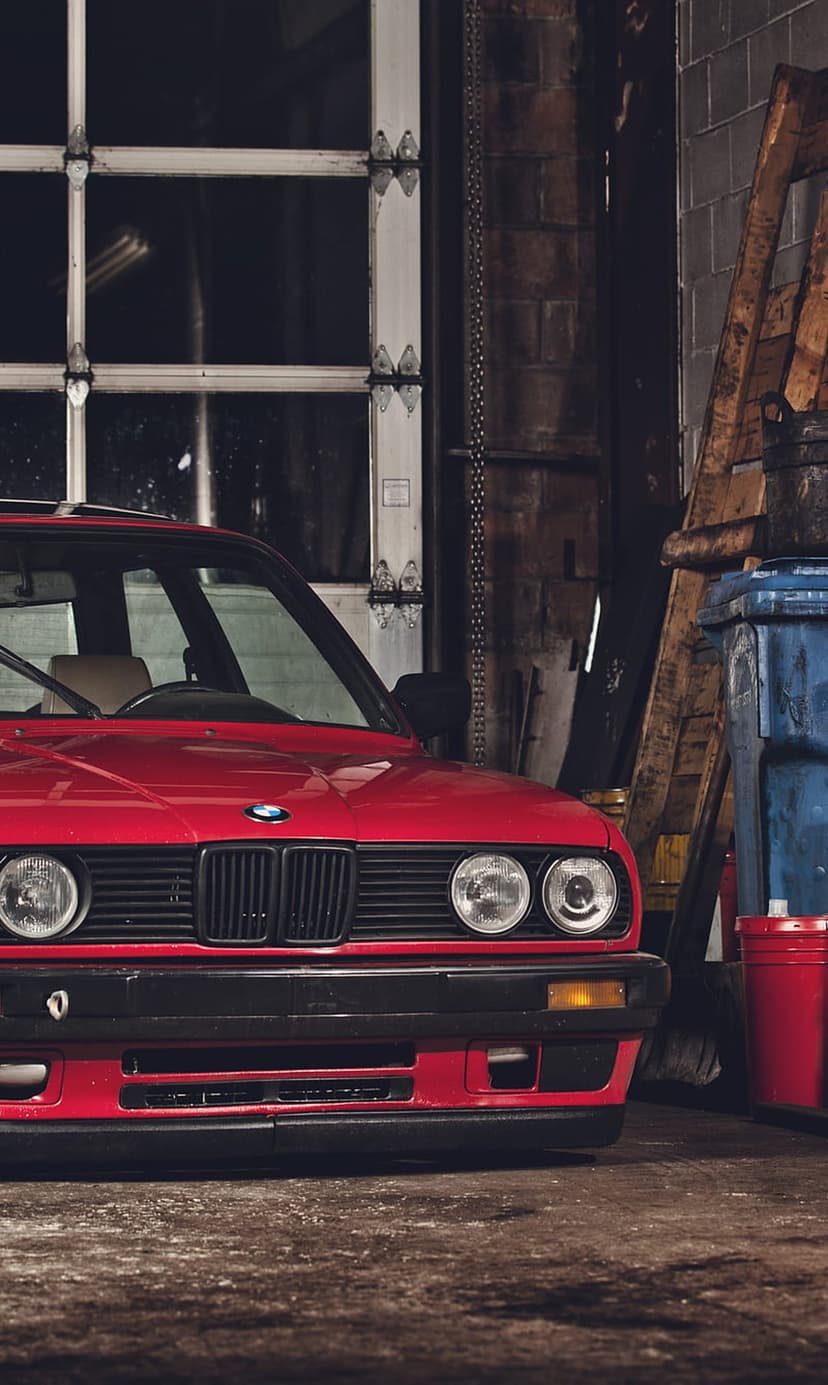Red BMW E30 sedan with round headlights and kidney grille parked in a dimly lit brick garage with metal doors.